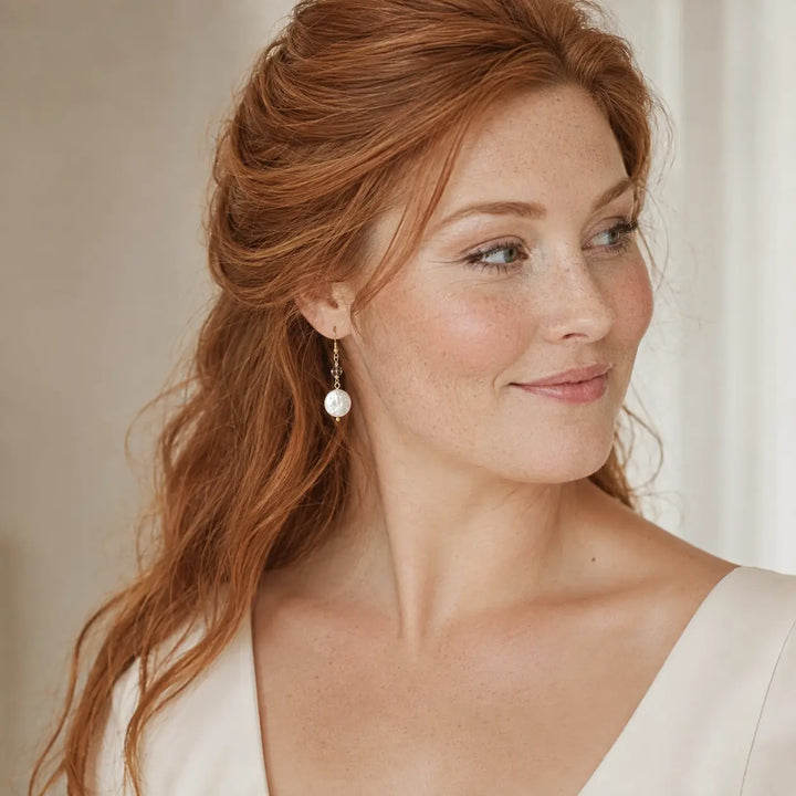A close-up portrait of a woman with red hair wearing a freshwater pearl drop earring with a gold-toned setting, against a soft neutral background.