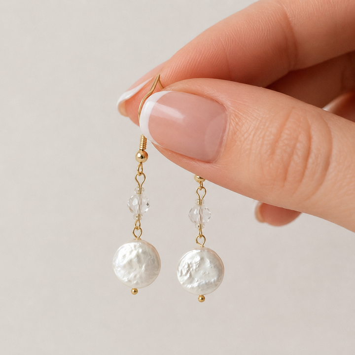 A close-up of a hand with a French manicure holding a pair of gold drop earrings with textured freshwater pearls and small clear beads, set against a soft beige background.