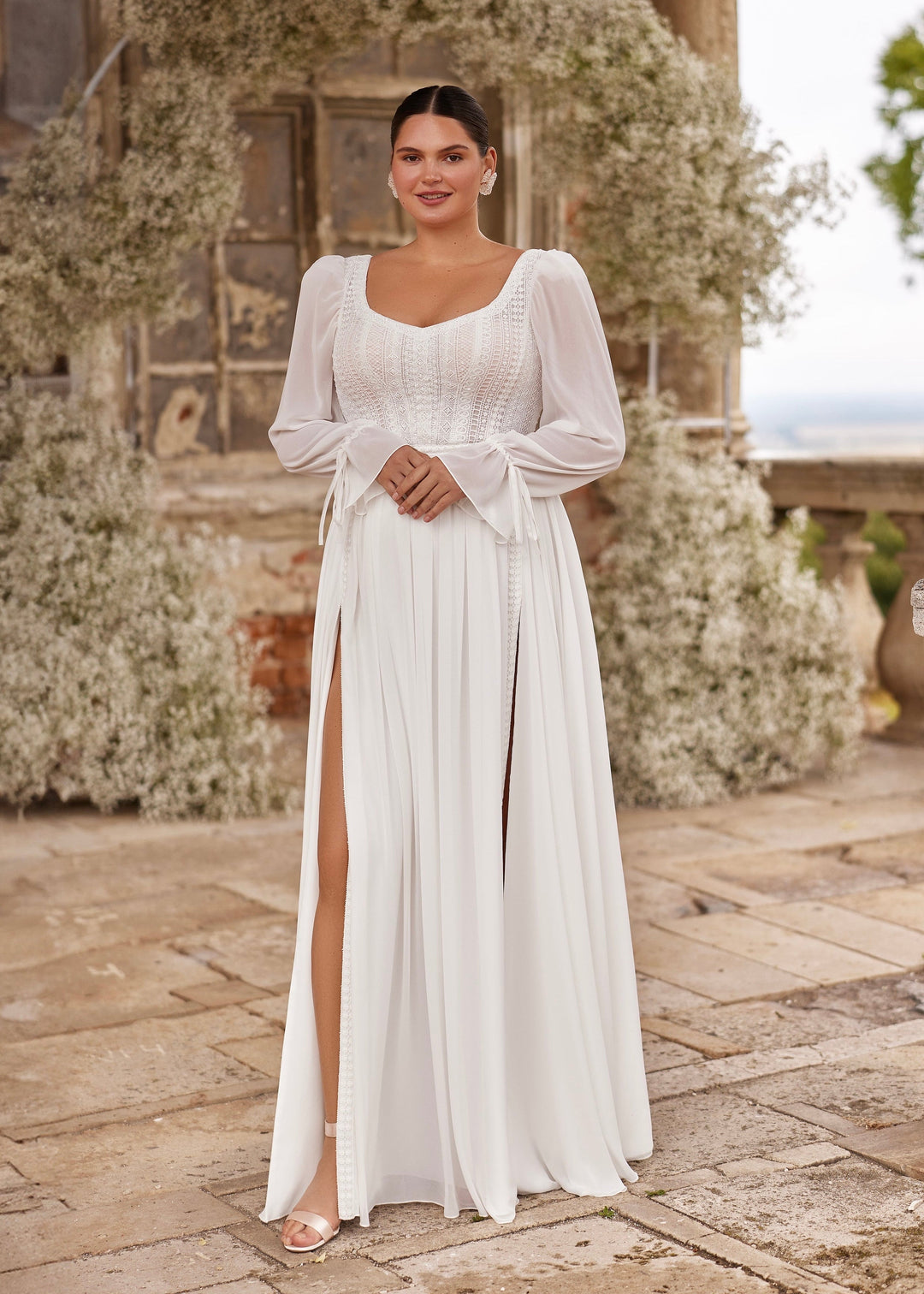 A woman wearing a white bridal gown with a lace corset bodice, long sleeves, and a chiffon skirt with front leg splits. The dress has a trainless design.