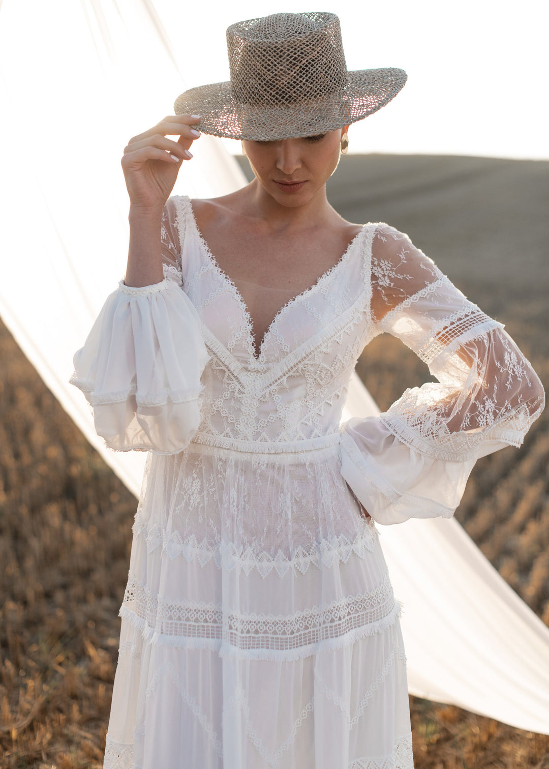 Woman wearing a boho lace a-line wedding dress and straw hat in a field setting.