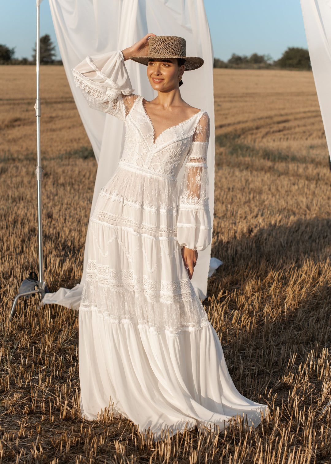 Woman in a boho chiffon wedding dress and hat standing in a field with a white curtain.