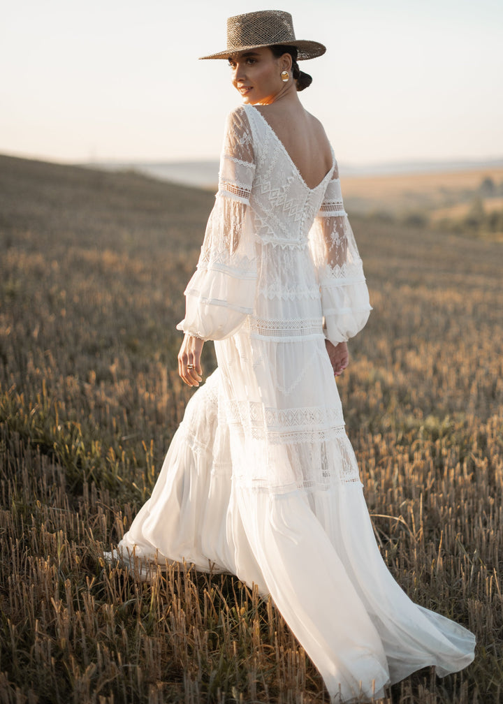 Woman in a boho a-line wedding dress and hat standing in a field with a scenic background.