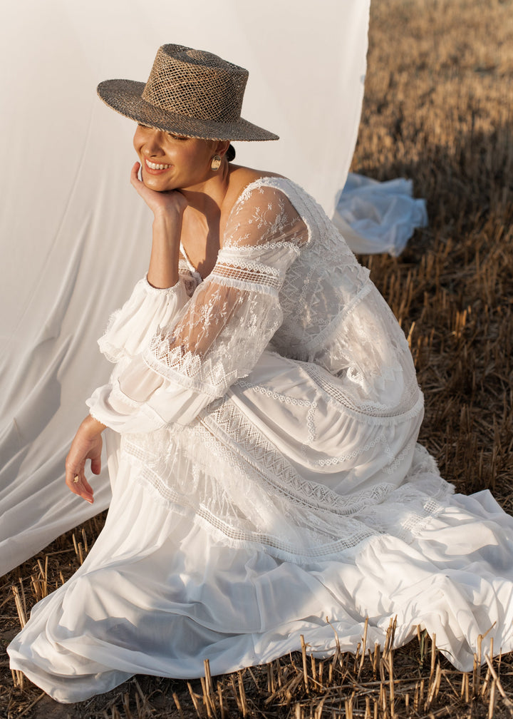 Woman in a boho wedding dress and straw hat sitting in a field with a white curtain in the background
