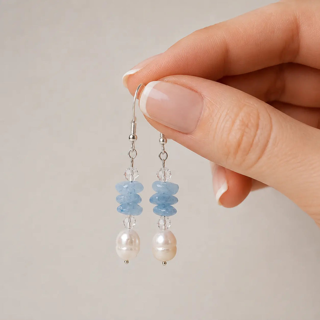 A close-up of a hand holding a pair of drop earrings against a soft neutral background. The earrings feature light blue stones, clear faceted crystal, and freshwater pearls on silver hooks.