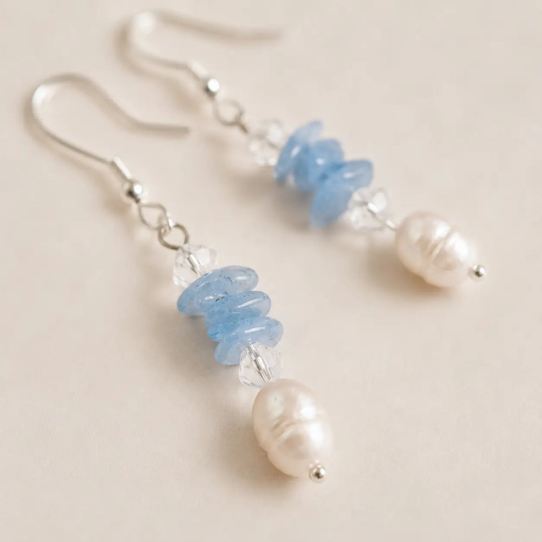 A pair of dangle earrings with silver fishhook wires, clear faceted beads, light blue stones, and freshwater pearls, displayed on a light beige background.