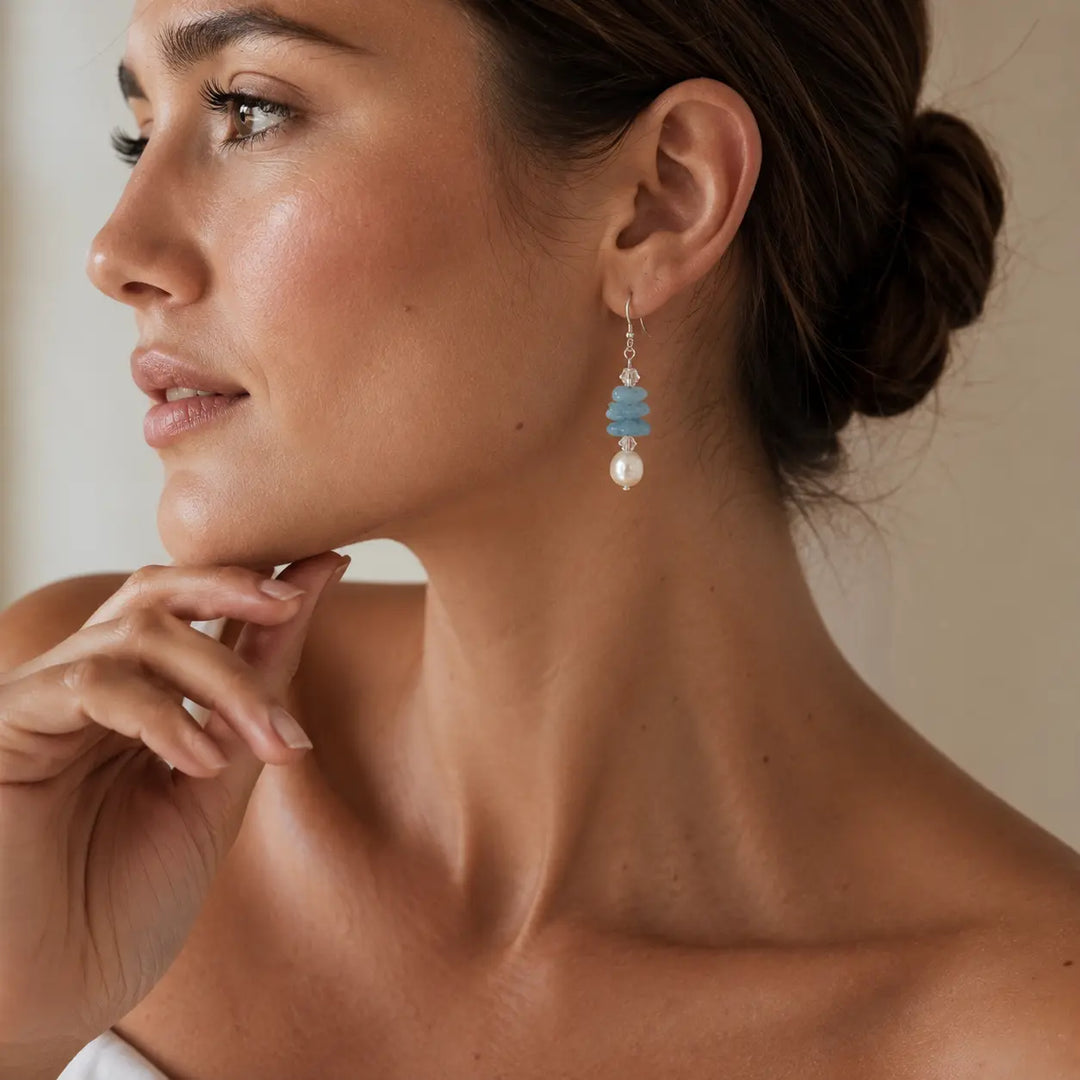 A close-up of a woman wearing a single drop earring with stacked light blue stones, a silver-toned metal connector, and a freshwater pearl dangle, shown against a soft beige background.