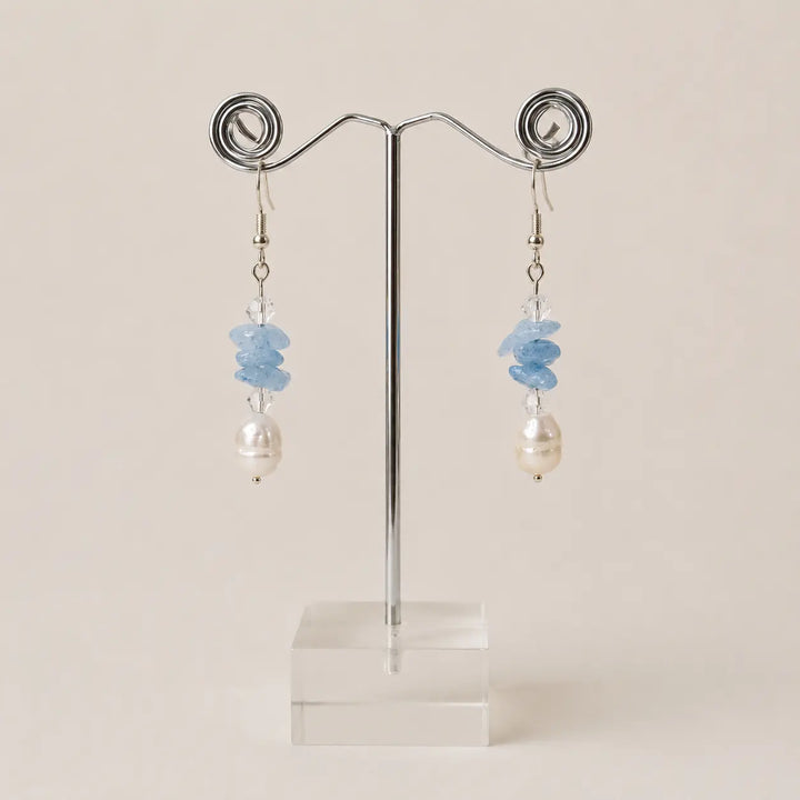 A pair of drop earrings displayed on a metal stand against a plain light beige background. Each earring has a cluster of light blue stones above faceted clear crystals and freshwater pearls.