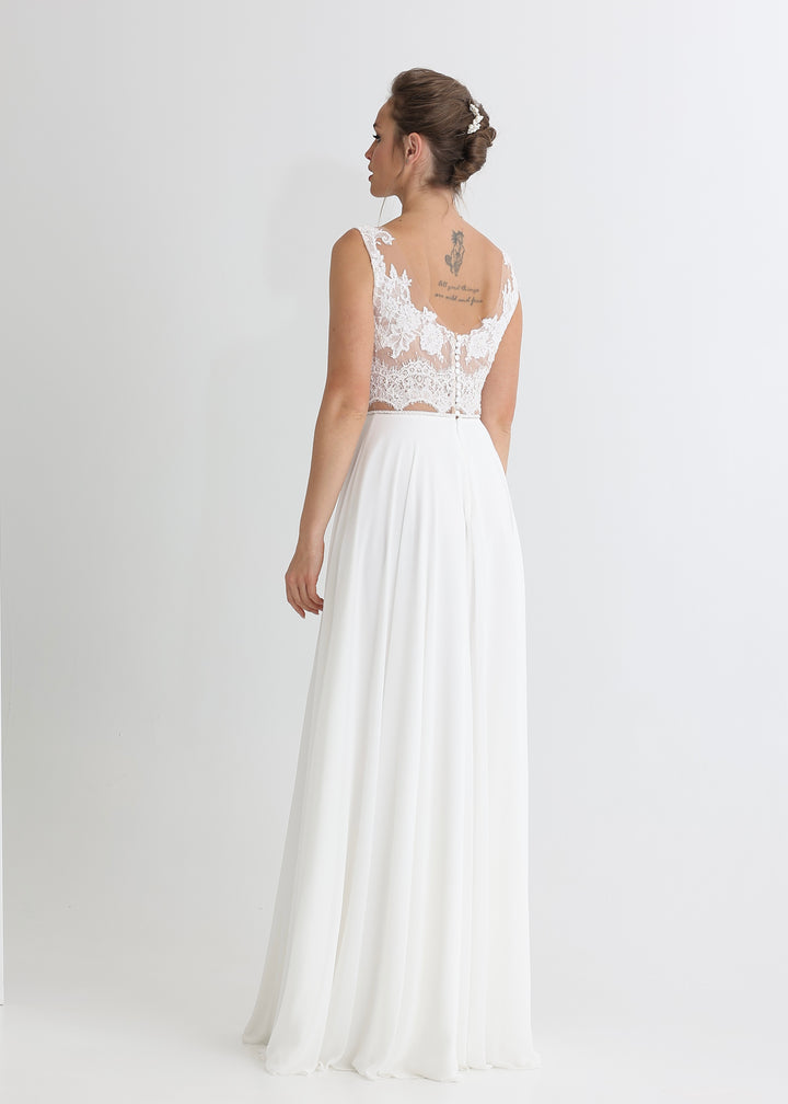 A woman wearing a white wedding dress with a lace top and chiffon a-line skirt stands with her back to the camera against a white baground.