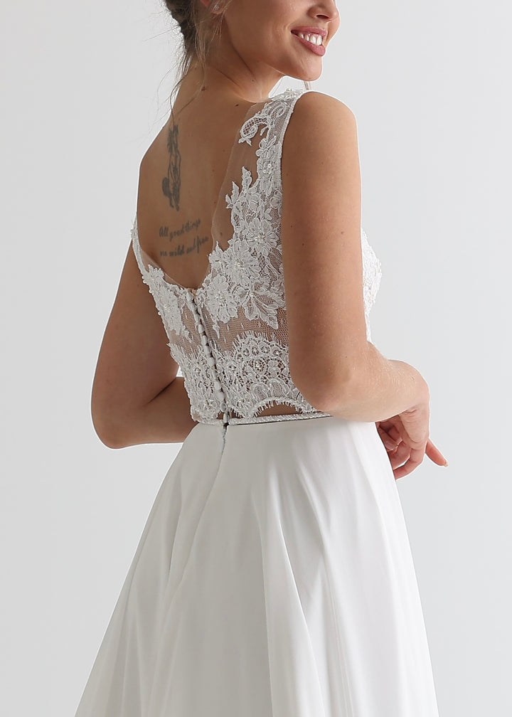 A woman standing with her back to the camera wearing a white wedding dress with a see-through lace top and simple chiffon, a-line skirt in a white studio background.