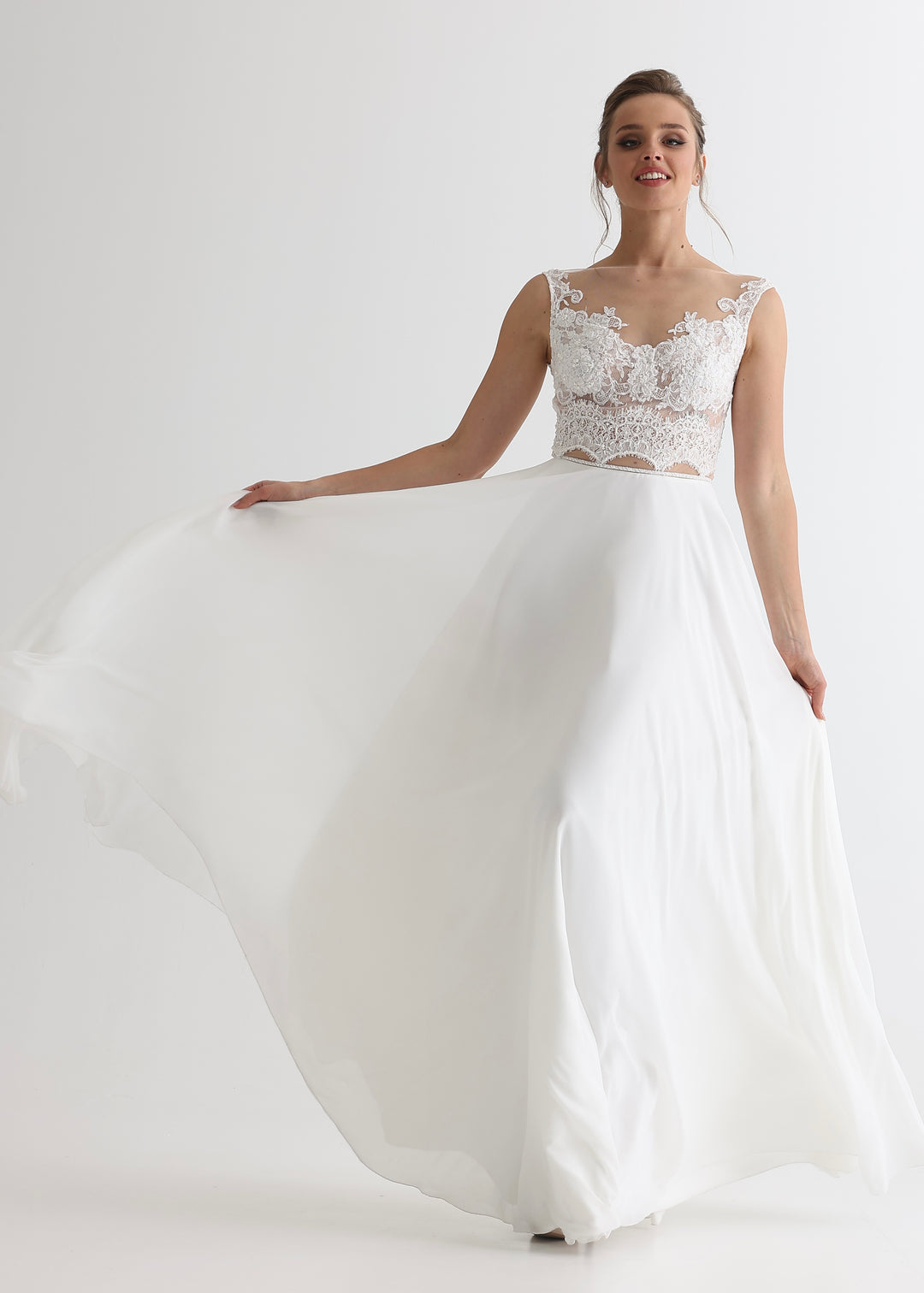 A woman wearing a white wedding dress with a sheer lace top and simple a-line skirt standing in front of a white studio background.