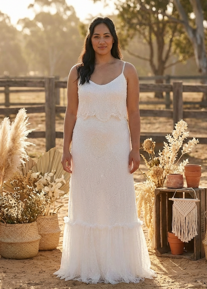 Woman in a beaded white lace dress standing outdoors with rustic decor.